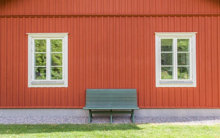 Röd träpanelvägg med två vita fönster ovanför grundsockel och grön bänk framför huset.