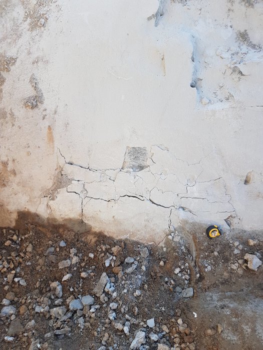 Betongvägg med sprickor och flagning vid grunden med mätkapsel bredvid grus och småsten.