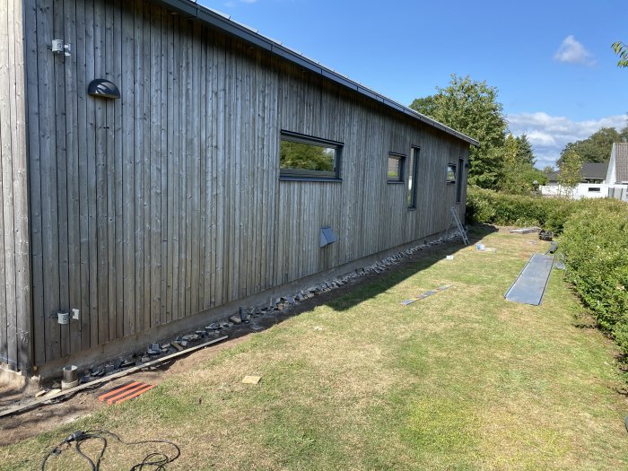 Husvägg med träpanel där underkanten precis har blivit skuren, med lösa panelbitar och verktyg på marken.