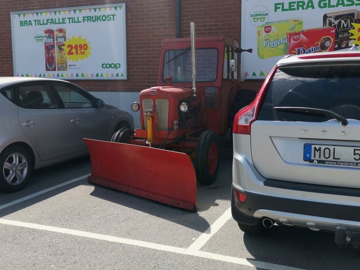 Röd traktor med snöblad parkerad på en matbutiksparkering mellan personbilar.