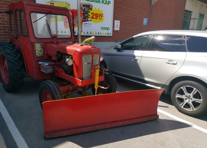 Röd 1961 T320 traktor med snöplog framför Konsum, parkerad bredvid modern bil.