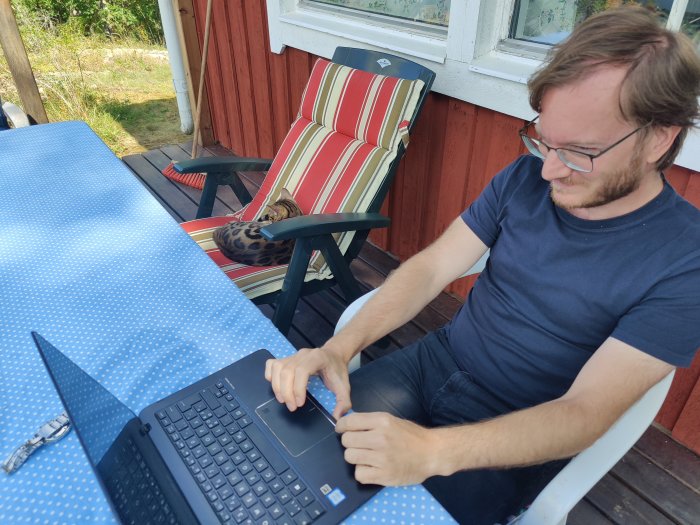 Man arbetar på laptop vid utomhusbord medan katt sover i bakgrunds solstol.