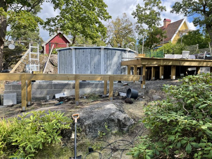 Stomme av ett nybyggt trädäck på bergig tomt, byggmaterial och verktyg syns med hus i bakgrunden.