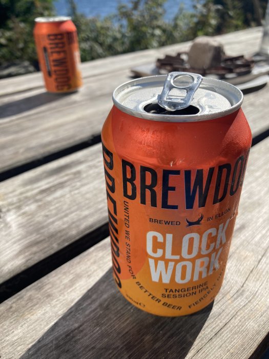 Öppnad ölburk av märket BrewDog Clockwork på ett träbord utomhus, med en oöppnad burk i bakgrunden.