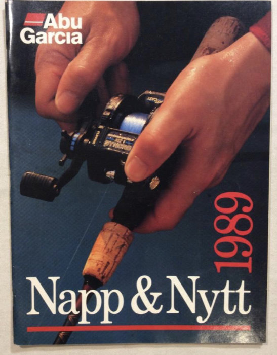 Hand håller en Abu Garcia fiskerulle över en katalog med texten "Napp & Nytt 1980".