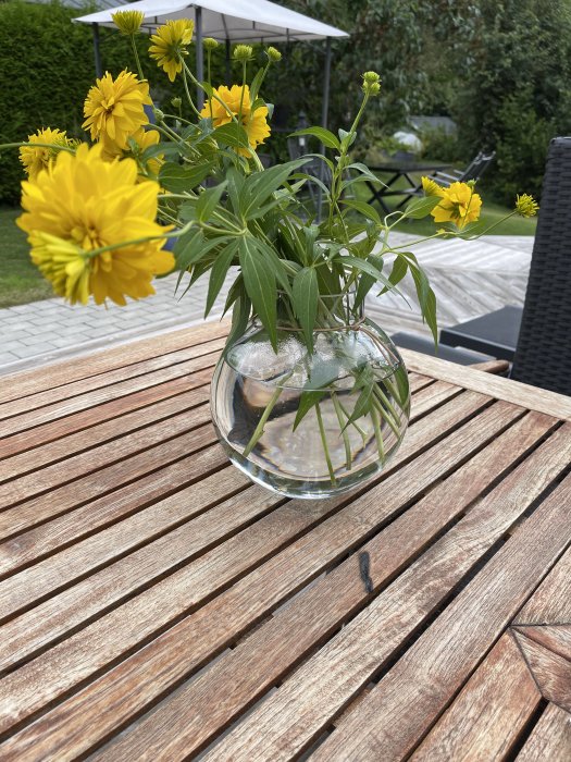 En bukett gula blommor i en genomskinlig vas på ett brunmönstrat teakbord med synligt jack.
