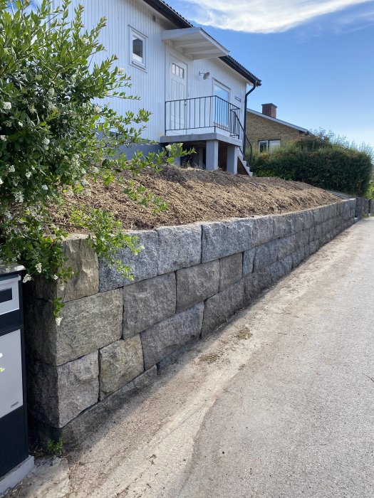 Stödmur av sten framför ett hus med sprängsten bakom och ett tunt jordlager ovanpå.