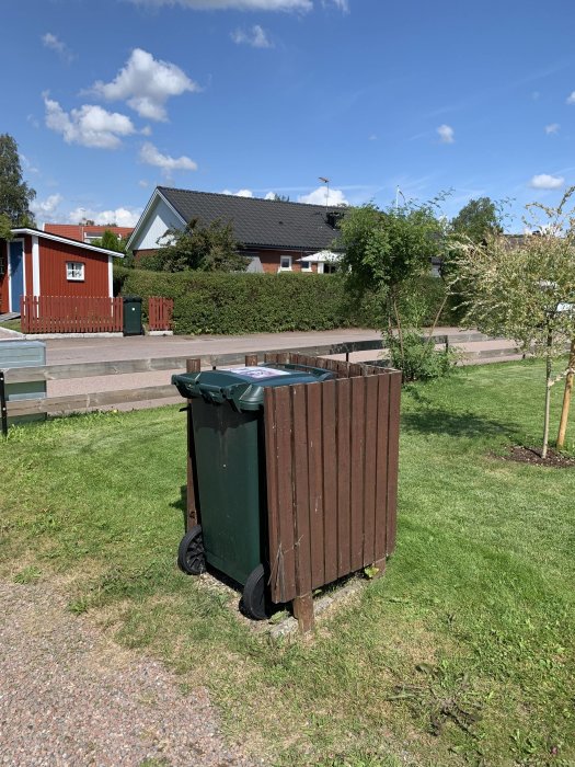 Soptunna skyddad av ett trästaket utan tak i en trädgård, hus i bakgrunden.