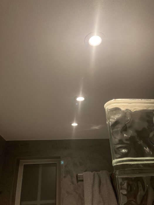 Inbyggda spotlights i ett mörkt tak med en glasblocksvägg och handdukshängare i förgrunden.