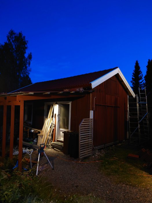 Renovering av röd stuga vid skymning med pågående takarbete och byggmaterial synligt.