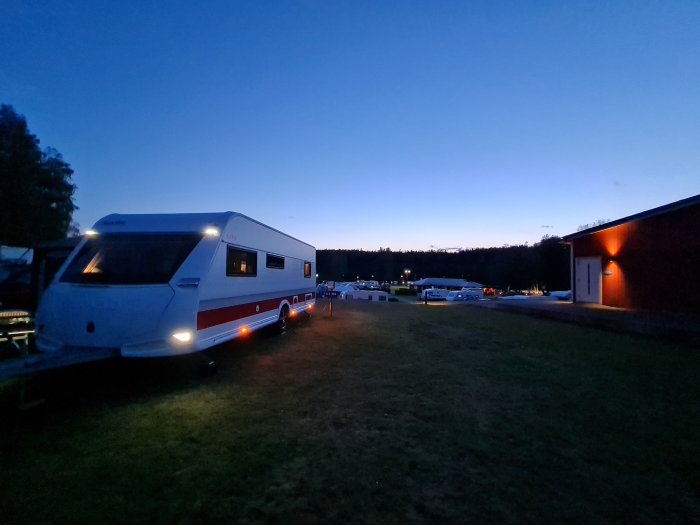 Husvagn parkerad på campingplats vid skymning med tända positionsljus och svagt ljus på himlen.