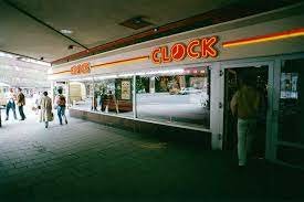 Exteriör av "CLOCK" snabbmatsrestaurang med neonskylt och folk som passerar framför.