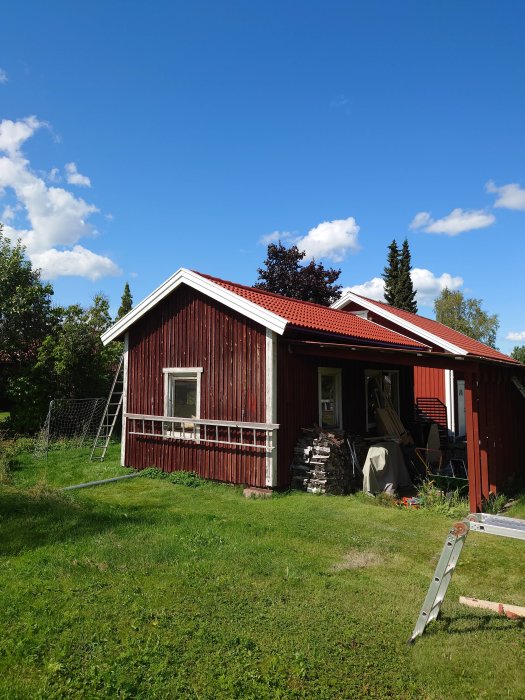 Röd stuga med nytt tak av pannor, plåt och ledig stege, arbete pågår, i soligt väder.