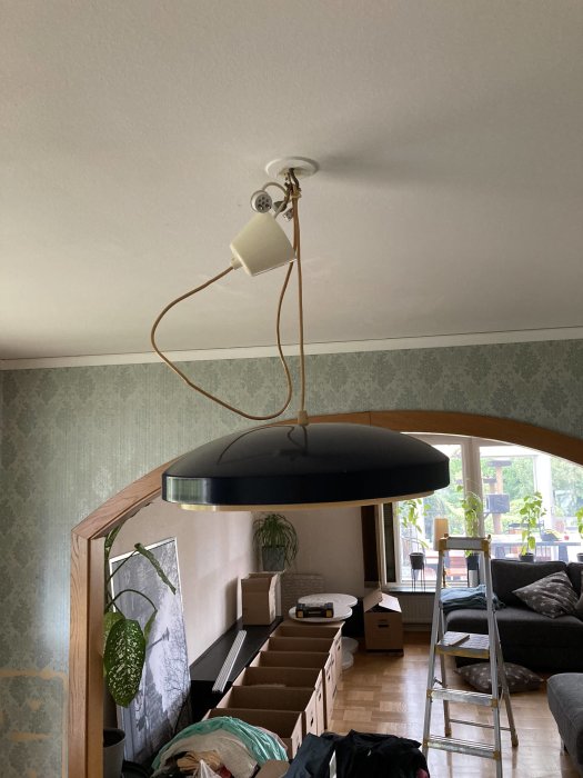 Taklampa av märket Belid, stor svart skärm och böjd guldfärgad stång, i ett rum under renovering.