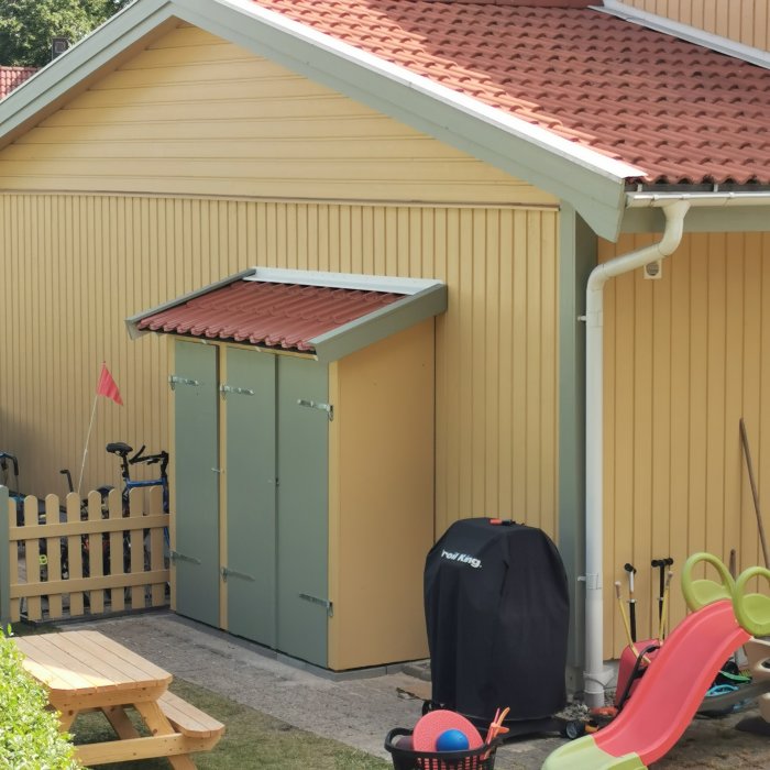 Nybyggt litet förråd vid sidan av ett gult garage, med tegelröd dörr och tak.