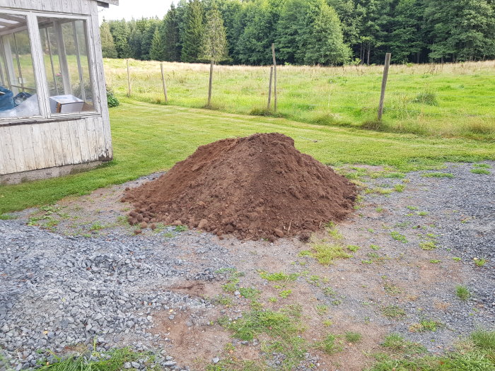 Ny jordhög bredvid grusparkering och grönt fält, nära en byggnad med skog i bakgrunden.