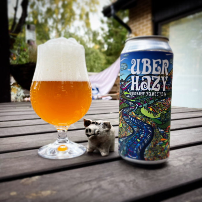 Ölglas med skum på träbord bredvid burk av 'Über Hazy' och en liten griskeramik.