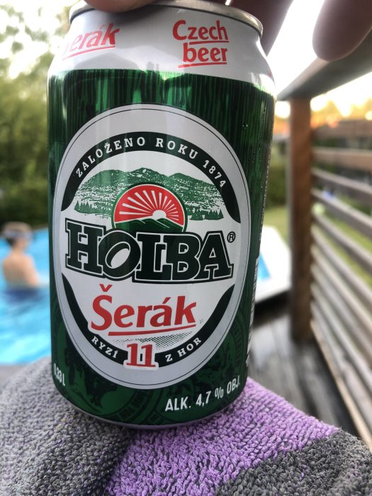 En hand håller en Holba Šerák tjeckisk ölburk mot en bakgrund av en altan och en pool.