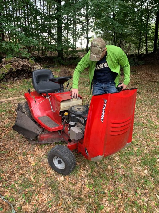 Person i grönt och mössa kontrollerar motor på röd äldre åkgräsklippare i skogsmiljö.