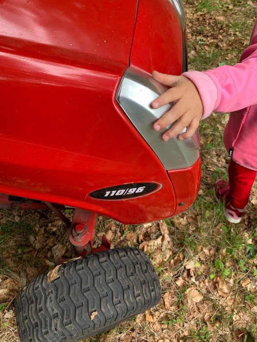 Barnhand på röd gräsklippare modell 110/96, delvis synlig däck och skogsmark i bakgrunden.