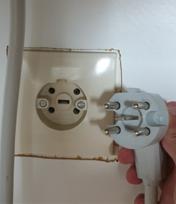 Hand håller en perilaxkontakt med tre stift nära en väggmonterad socket i en köksmiljö.