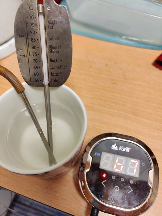 Analog köttermometer i kopp med vatten jämförs med digital iGrill termometer som visar 67.7 grader.