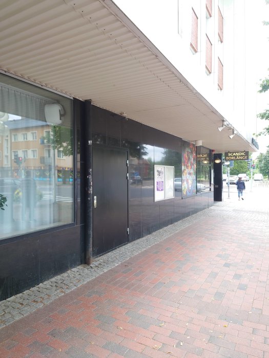 Svart anonym dörr vid trottoaren nedanför Scandic Borlänge, med reflektioner i fönstret.