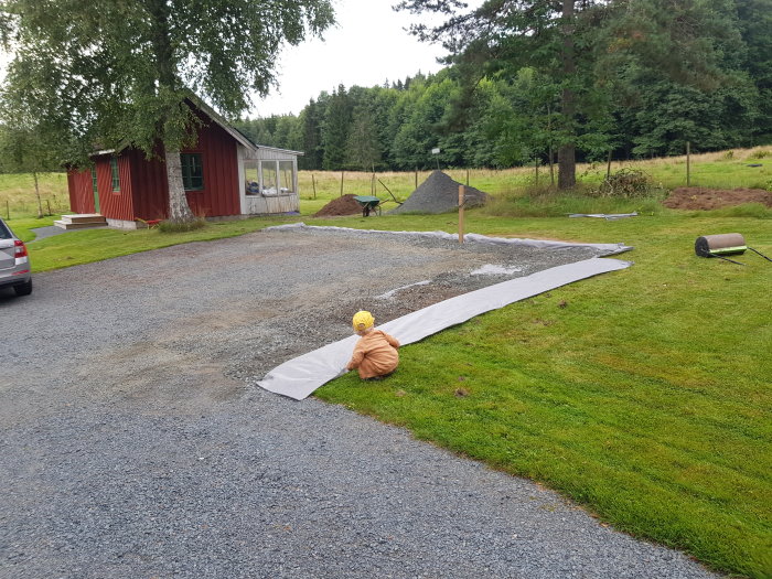 Person rullar ut markduk på grusunderlag nära röd stuga och bil, med gräsmatta och trädgårdsarbeteverktyg synliga.