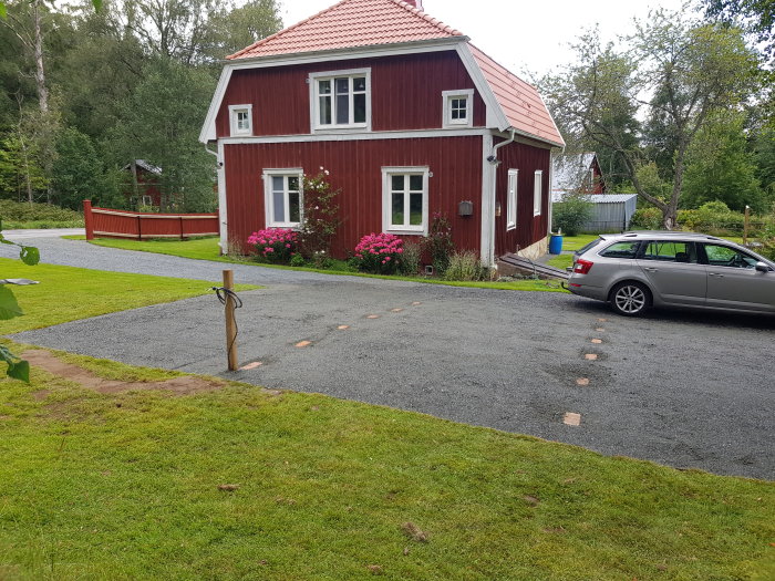 Rödfärgat hus med nyanlagd grusgång och nysatta plantor, grön gräsmatta och bil parkerad på gården.