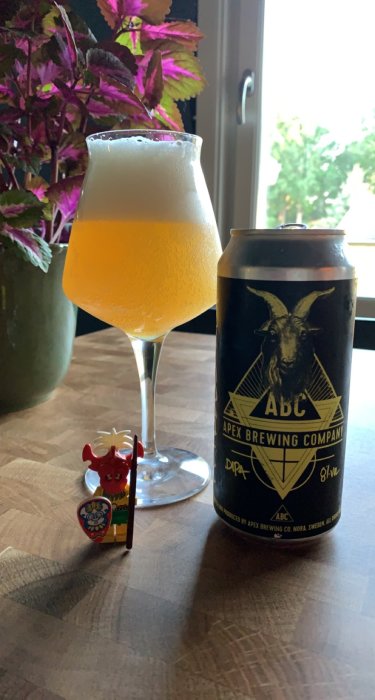 En ölglas fyllt med öl bredvid en ölburk från Apex Brewing Company och en leksaksfigur på ett bord.