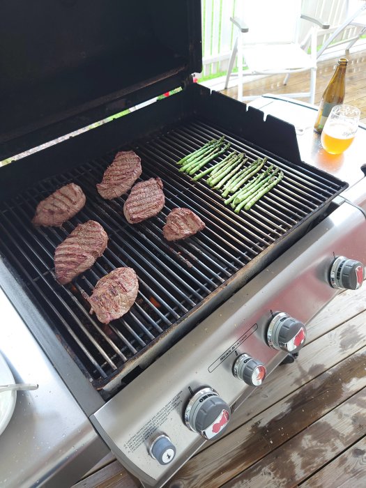 Grillat kött och sparris på en gasolgrill med en öl bredvid på en träterrass.