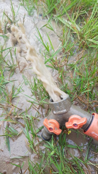 Vatten pumpas ut genom en grå och orange Gardena koppling på lerig mark med gräs.