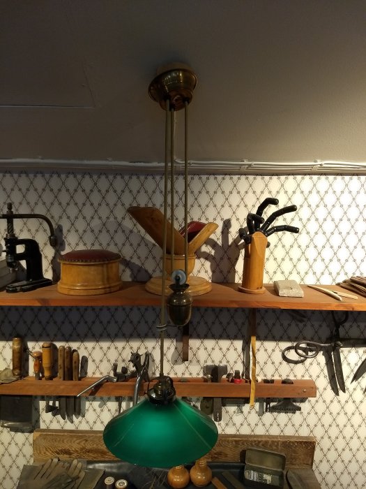 Gammal justerbar taklampa med grönt glas och mässingdetaljer på ett museum med verktyg i bakgrunden.