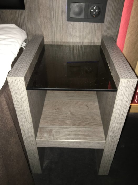 Mörkt träliknande nattduksbord intill en säng med trätexturerad yta och en spegelglas-överdel.