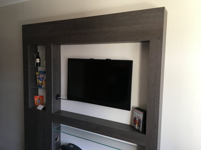 Mörkbrun träliknande mediavägg med inbyggd TV, hyllor och dekorativa föremål i ett hotellrum.