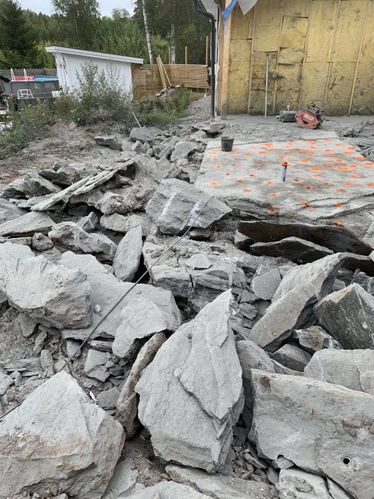 Sprängningsarbete i berggrund med synliga borrade hål och oranga markörer mot bakgrund av byggarbetsplats.