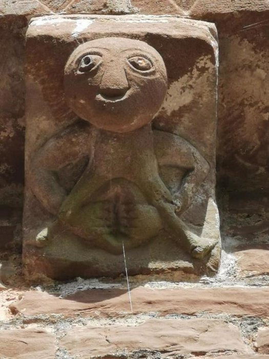 Skulptur i sten som föreställer en sittande figur med ett leende ansikte, inmurad i en tegelvägg.