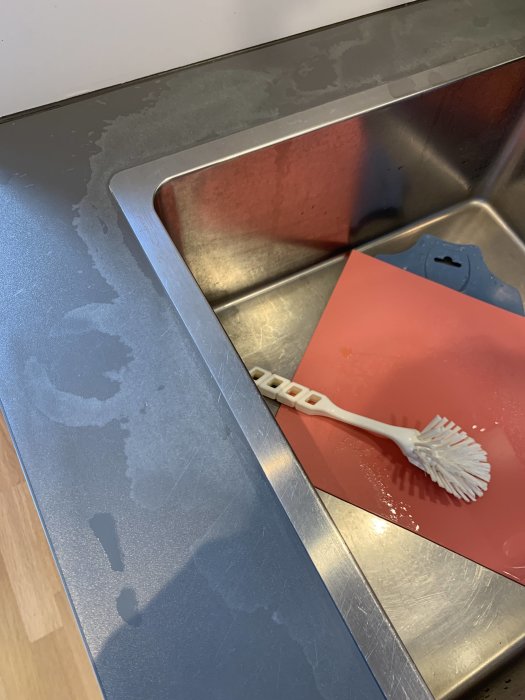 Bänkskiva med blekta fläckar vid diskho efter spill av kalklösningsmedel, diskborste och röd matta synlig.
