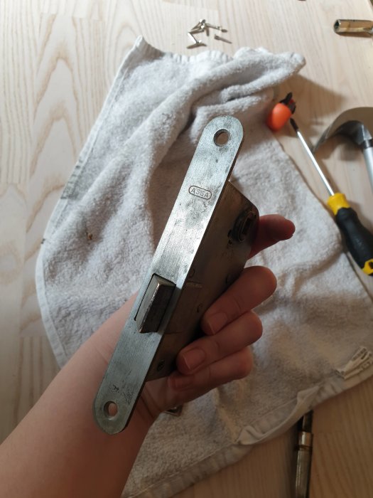 Hand håller en demonterad dörrlåsmekanism från ASSA, verktyg och handduk syns i bakgrunden.