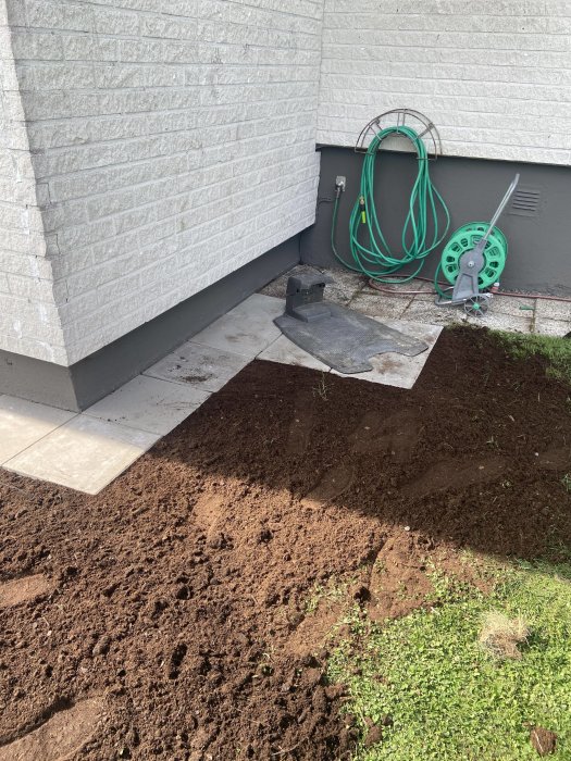 Nyinstallerade plattor vid husvägg med fogsand, utlagd jord och gräsfrö samt en laddstation och slangvinda.