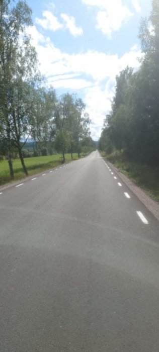 Asfalterad landsväg omgiven av träd under en solig dag med blå himmel och vita moln.