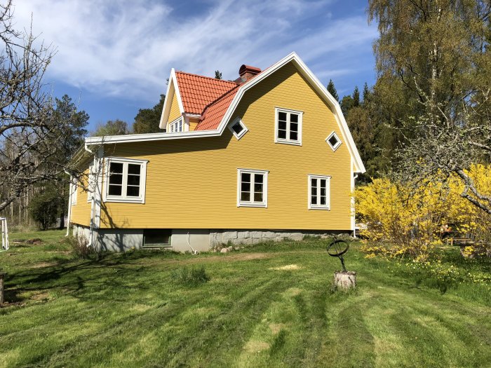 Gult hus med torpargrund och tillbyggnad, omgiven av grönska och en blommande buske.