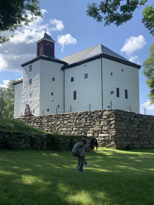 Två personer leker framför en stor vit byggnad med rött tak och klocktorn, omgiven av stenmur och grönt gräs.