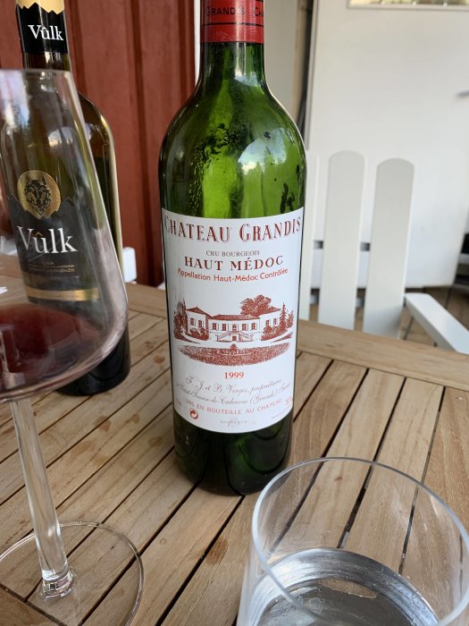 Flaska Château Grandis Bordeaux och Vûlk vin på ett träbord med ett vinglas i förgrunden.