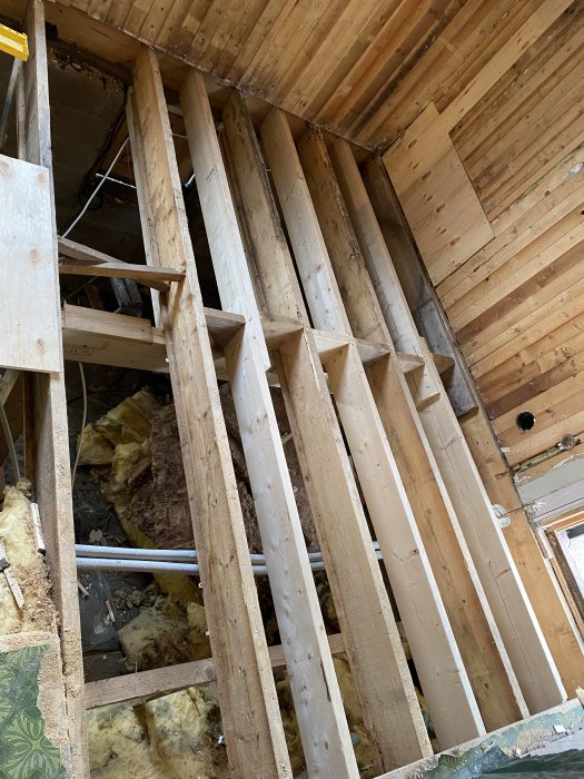 Reglar överlappar varandra i en oisolerad vägg under renovering, med synlig isolering och träplank.