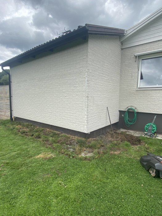 Hörn av hus med vitputsad fasad, nyinstallation av stuprör och nylagd dränering mot gräsmatta.