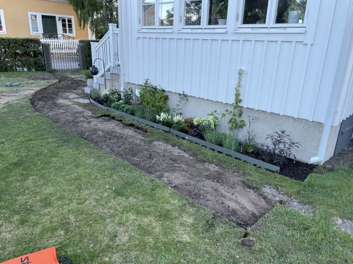 Nyplanterad rabatt längs med husväggen och utgrävda konturer för grusgångar i trädgården.