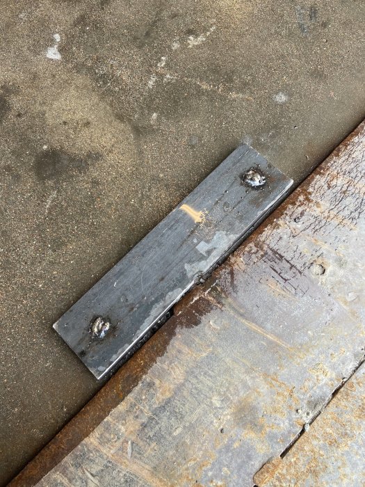Ett metallstycke med amatörmässigt utförda svetsfogar placerat på en rostig arbetsyta.