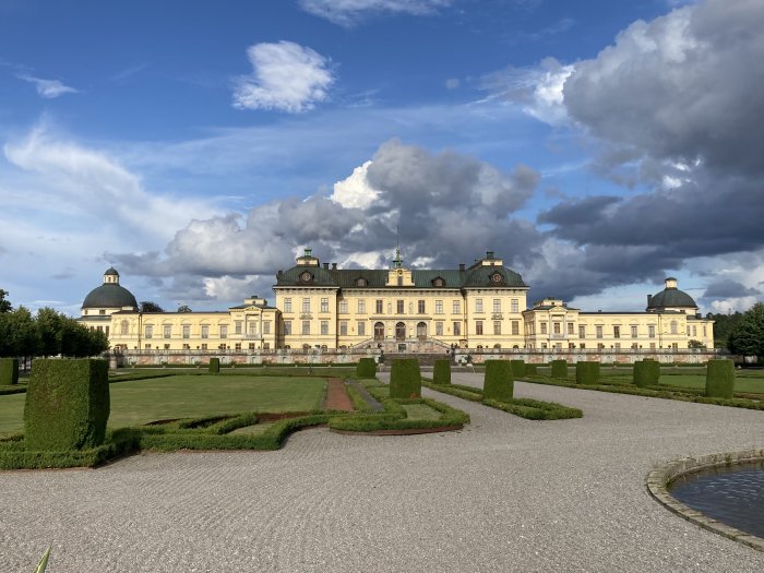 Ståtlig klassisk slottsbyggnad med kupoltak omgiven av välskötta gröna trädgårdar och promenadvägar under en molnig himmel.