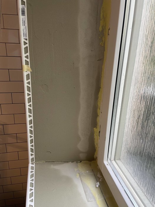 Halvfärdigt badrumsrenovering med brun kakling, ojämn gipsvägg och uncementerat golv nära ett fönster.